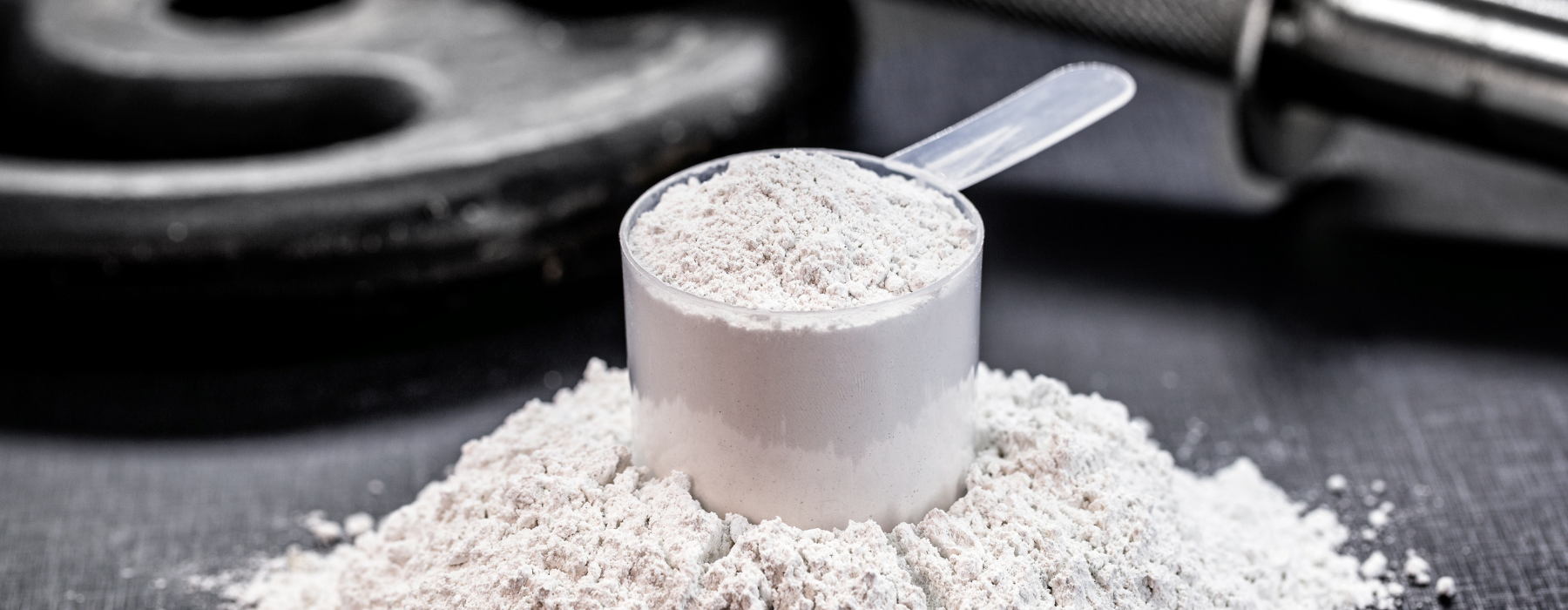Medidor transparente cheio de creatina em pó sobre uma superfície escura, com pesos de academia desfocados ao fundo.