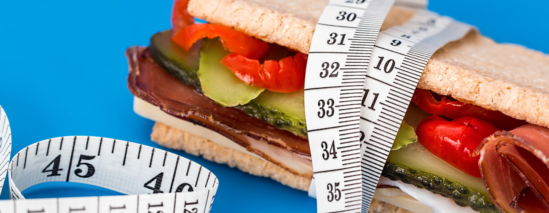 Sanduíche envolto por fita métrica sobre fundo azul, simbolizando o controle do peso, a alimentação e a dificuldade de emagrecer devido ao peso set point.