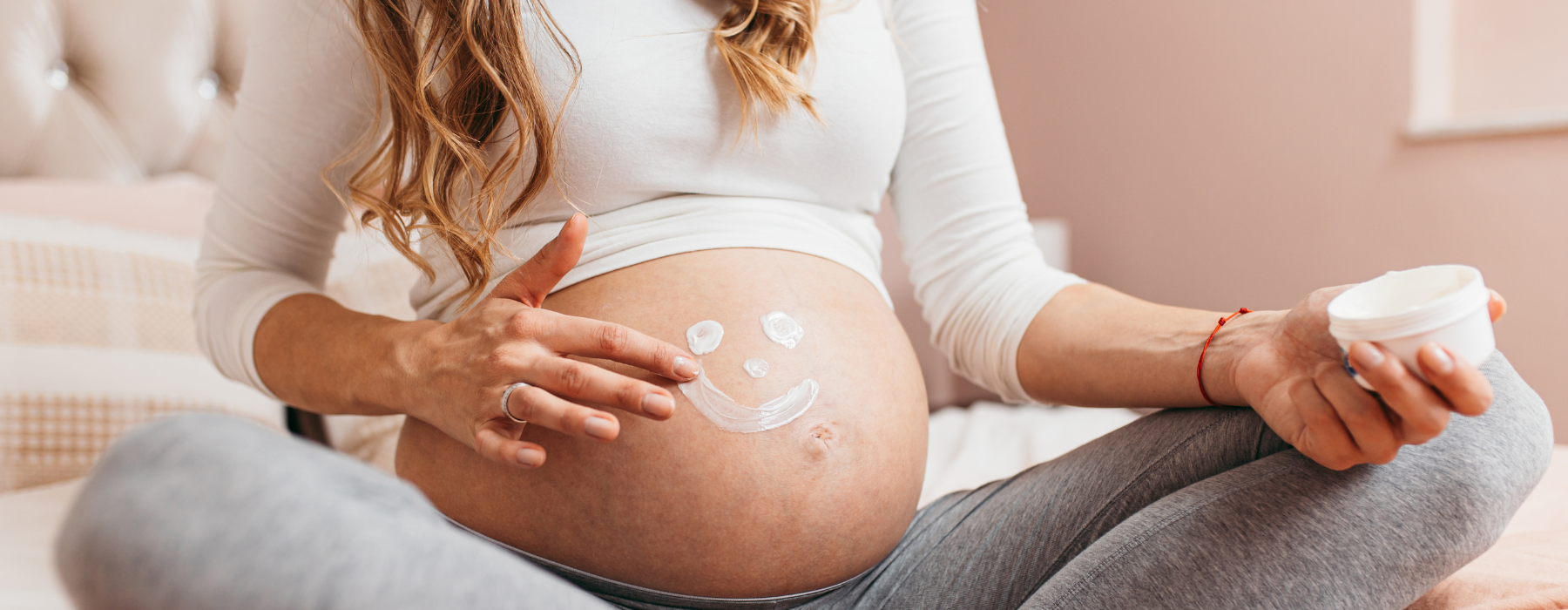 Mulher grávida sentada, com as mãos aplicando creme na barriga, desenhando um sorriso sobre a pele.