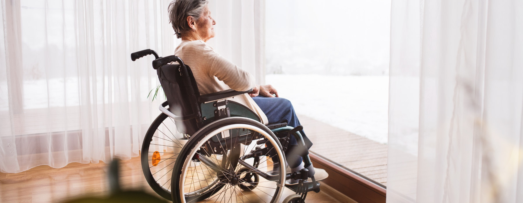 Mulher idosa sentada em cadeira de rodas próxima a uma janela, representando perda de mobilidade causada pela Esclerose Lateral Amiotrófica.
