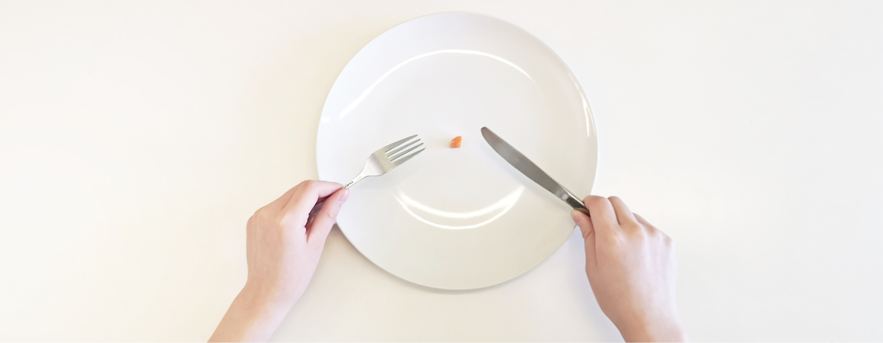 Mãos seguram garfo e faca diante de um prato quase vazio com um pequeno pedaço de comida, ilustrando a restrição alimentar relacionada à anorexia nervosa.